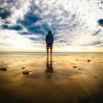 A lone figure stands on a tranquil beach during a vivid sunset, reflecting on the wet sand.